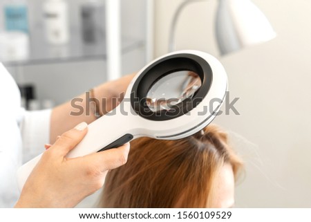 A trichologist examines the condition of the hair on the patient’s head with a dermatoscope. In a bright cosmetology room.
