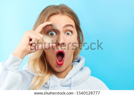 Young beautiful woman looking at the camera through magnifier with funny surprised expression, closeup isolated on blue. Enlarged eye, optical illusion, search