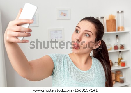 Young chubby woman standing in kitchen taking selfie photo on smartphone showing tongue to camera smiling playful close-up