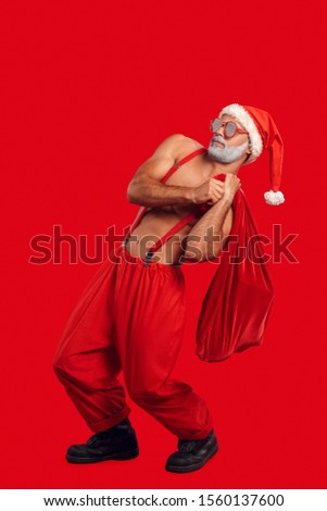 Young bearded Santa Claus bare muscular upper body wearing hat and sunglasses standing isolated on red background christmas concept holding heavy gift bag leaning back side view