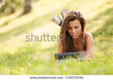Pretty brunette girl is looking a digital screen while sitting on the grass and smiling