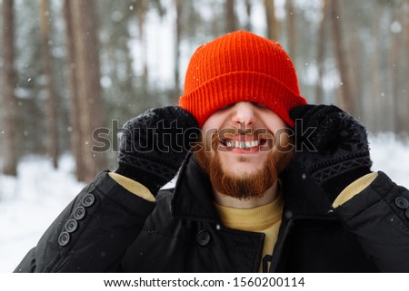 Portrait of a charismatic guy with a beard in a red hat and winter stylish clothes in the style of casual in a winter snowy forest