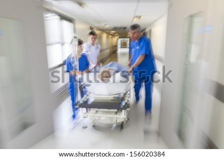 A motion blurred photograph of a patient on stretcher or gurney being pushed at speed through a hospital corridor by doctors & nurses to an emergency room Royalty-Free Stock Photo #156020384