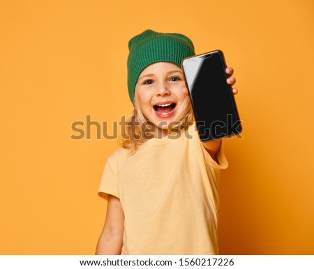 Smiling little girl kid in green modern winter hat showing blank screen of new popular mobile phone on light yellow background