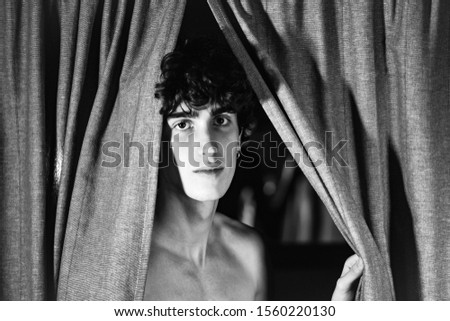 black and white portrait photography. expressive and attractive handsome young teenager Italian model boy posing for fashion shooting and hiding behind curtains