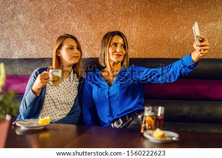 Two women adult female friends or sisters sitting in cafe taking self portrait photos selfies using mobile smart phone smartphone