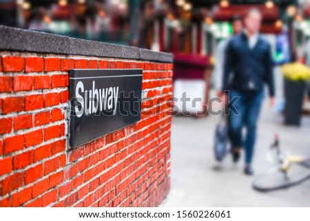 New York City street corner subway entrance with sign on brick wall and blur of men walking in the background