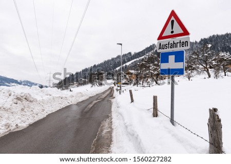 Red triangle road traffic warning sign - Other Danger with text Ausfahrten which means Exits. Snow covered road, trees and mountains. Austria