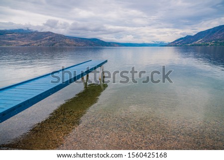 Blue painted dock extending over calm lake with view of overcast sky and mountains
