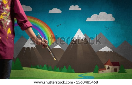 Close up artist hand holding paintbrush. Painter in shirt standing on background colorful picture. Summer landscape with mountains, blue sky and rainbow artwork. Creative hobby and profession.