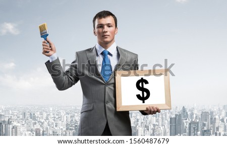 Creative businessman painter holding paint brush and whiteboard in hands. Portrait of young handsome man in business suit and tie on cityscape background. Ambitions and creativity in business.