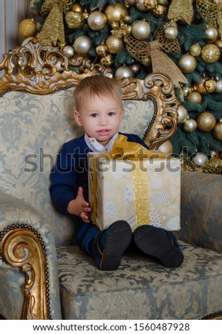 Happy little boy sitting by the Christmas tree in a blue suit with a sweater. child with a gift in a golden box.