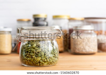 Beautiful closeup shot of a glass jar airtight container full of dried pumpkin seeds or pepita on the wooden kitchen counter. The seeds contain lots of nutrients benefit for heart health and bones. Royalty-Free Stock Photo #1560602870