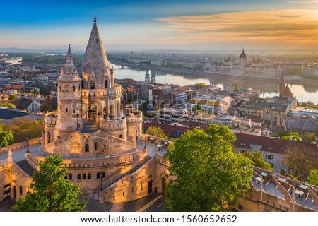 Budapest, Hungary - Beautiful golden summer sunrise with the tower of Fisherman's Bastion and green trees. Parliament of Hungary and River Danube at background. Blue sky. Royalty-Free Stock Photo #1560652652