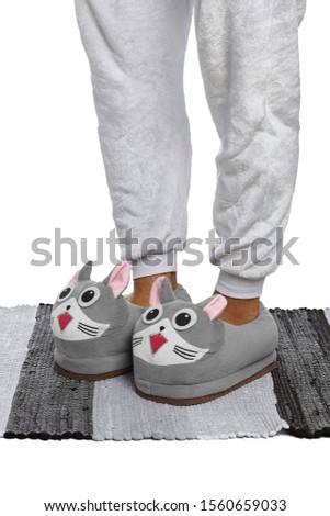 Close-up shot of male legs in white velour trousers and gray plush house slippers made in the form of kawaii cat. The man is standing on the striped gray and white carpet.