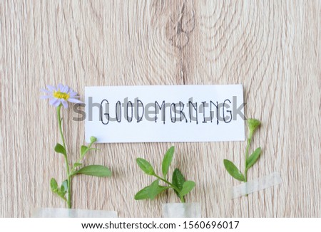 flower  and  green leaf stick by paper tape on wooden  background with words " good morning " written by hand on a piece of white paper 