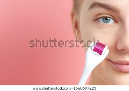 Close-up female face with dermaroller for mesotherapy procedures, skin care at home and in salon. Meso roller with microneedles on pink background Royalty-Free Stock Photo #1560697223