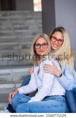 A beautiful blonde with glasses and her mature mother are sitting on the stairs outdoor. A well-groomed elderly woman hugs her adult daughter tightly. Love and family values. Mothers Day.