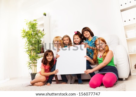 Large group of happy kids sitting in the sofa in domestic room holding blank advertising board.