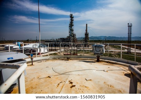 Grey distillation column under bright sun. Blue sky with clouds and mountains background. Oil refinery and gas processing plant in desert. Yellow oil storage tank on foreground. Taraz city.