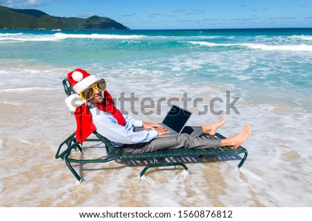 Relaxed businessman in Santa hat, sunglasses and big red Christmas bow working on his laptop in the waves of a tropical Caribbean beach