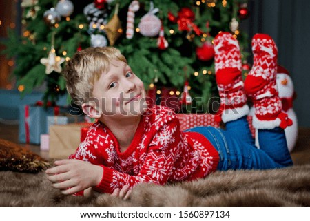 boy on Christmas eve under a Christmas tree with a gift box. Child opens Christmas gifts. Decorated living room with traditional red and green decoration. Cozy warm winter evening at home.