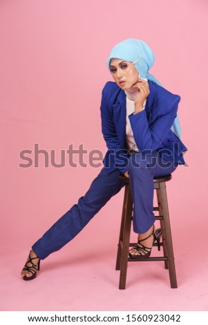 Beautiful elegance muslim lady , clothed in blue business suit, white cotton blouse with hijab sitting on a chair isolated over pink background.  Woman business corporate concept.