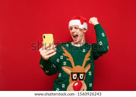 Portrait of a joyful guy in Christmas clothes on a red background, rejoices victory with a smartphone in his hand,looks at the screen and raises his hand with joy.Happy gamer is happy to win the game