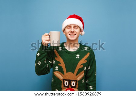 Portrait of smiling young man in green warm sweater and hat of santa claus, standing against blue background with cup hot drink in hand,looking into camera and smiling.New year and christmas concept