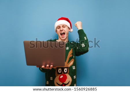 Portrait of joyful young man in green Christmas sweater and santa hat stands with laptop on blue background and rejoices in victory with raised hand. Happy guy with laptop rejoices. Isolated