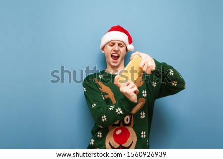 Expressive young man in santa claus hat and green christmas sweater plays video games on blue background, looks at smartphone screen and shouts. Emotional young gamer plays mobile games and Christmas