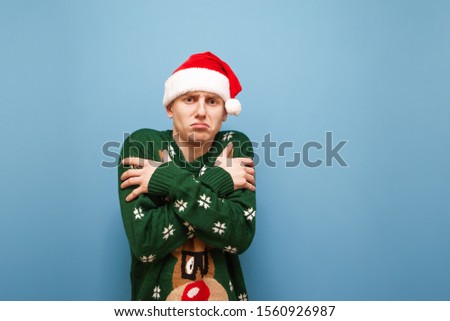 Sad young man in green warm sweater and santa hat warms up against blue background,guy freezes, looks in camera with sad face.Portrait of not cheerful guy froze in frost in winter, isolated. Christmas