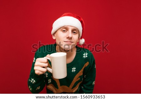 Portrait of a positive guy in Christmas clothes with a cup of hot drink in his hand stands on a red background, looks into the camera and smiles, wearing a Santa hat and a green warm sweater. Isolated