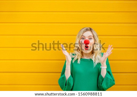 Funny young woman in green sweater red nose of a clown showing shock emotion on yellow background. Concept red nose day, holiday, party, clown concept. Royalty-Free Stock Photo #1560988811
