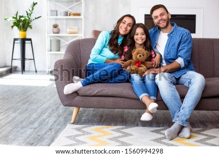 Our family portrait. Full-length family picture of mom, dad and their cute daughter with a teddy bear, smiling and hugging on the couch in the cozy home atmosphere.