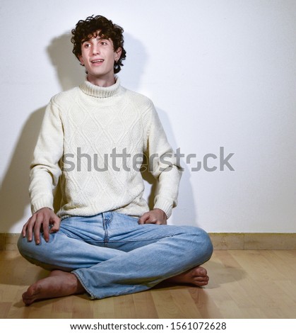 attractive and expressive young handsome teenager Italian model boy posing barefoot for a casual fashion shooting, sitting on wooden floor and wearing blue jeans and winter turtleneck