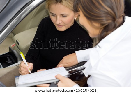 Woman sitting in the drivers seat signing for ownership of new car with the saleswoman pointing to the place to sign