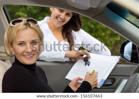 Happy beautiful blond woman signing for the purchase her new car with the smiling saleslady holding the contract through the open window