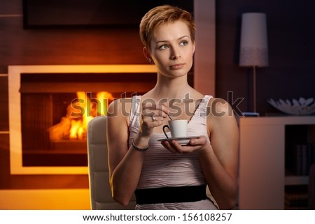 Daydreaming young woman drinking coffee at home by fireplace.