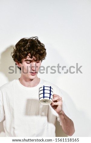 portrait of expressive young Italian teenager model boy posing for a fashion shooting, wearing a white shirt on white background and drinking a cup of tea/coffee