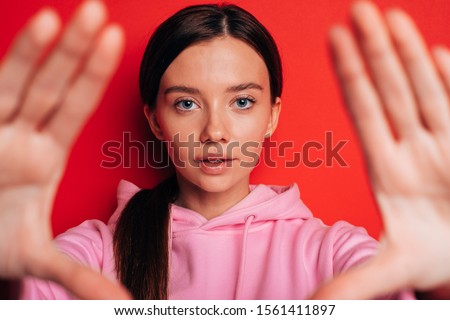 Calm peaceful young woman look on camera straight. Show hands close to shot. Wear pink hoody. Brunette isolated over red background