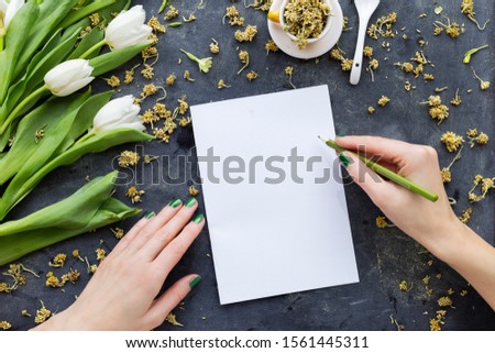 A person drawing on a white paper with a green pencil near white tulips with dry flowers on a black surface