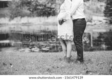 A grayscale of a pregnant couple standing o a grassy shore near a lake with their hands on the belly