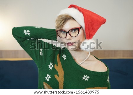 cute plus size nerd girl in glasses and green Christmas sweater with reindeer and red Santa Claus hat in the morning on the bed posing