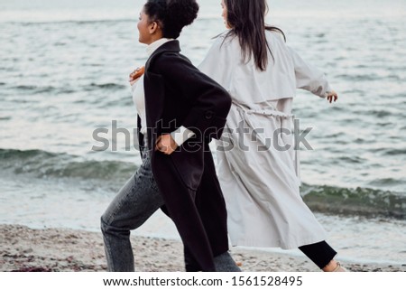 Two attractive stylish girls in trench coats happily running from back by the sea