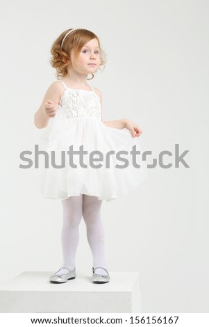 Little girl in white dress poses on big cube on white background.