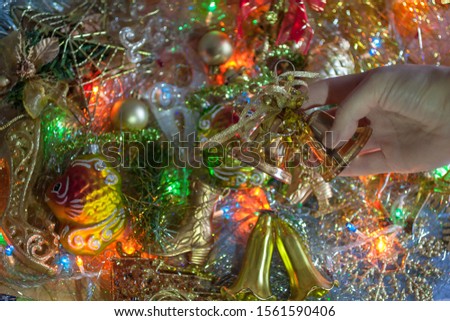 The female hand holds the Christmas bell on the others toys background (fish, deer, stars, skates, balls) with the silver tinsel and the colorful garland. The top view. New year/Christmas background