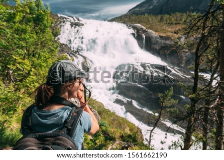 Kinsarvik, Hordaland, Norway. Young Woman Lady Tourist Traveler Taking Pictures Of Waterfall Nyastolfossen In Hardangervidda Mountain Plateau. Nyastolsfossen Is Famous Popular Destination.