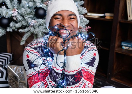 Portrait of positive african-american man in xmas sweater and warm white hat holding LED lights on background of Christmas artificial tree with black balls and gift boxes. Dark color picture