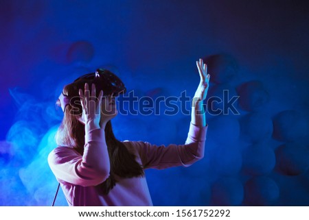 A delighted young woman in virtual reality glasses, against the background of a futuristic pattern on the wall in the shape of a pixel heart, get experience in virtual space.